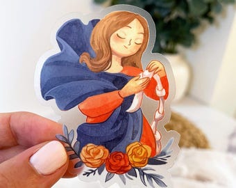 May include: A die-cut sticker featuring a watercolor illustration of a person with brown hair, wearing an orange top and a flowing dark blue shawl. The person holds a white ribbon, and the base is decorated with orange and yellow roses.