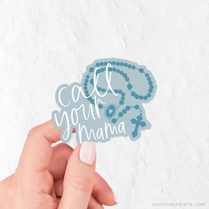 May include: A light blue sticker featuring a dark blue rosary illustration and the white cursive text call your mama. The sticker has a die-cut shape that follows the outline of the rosary beads and the cross, creating a cohesive graphic design.