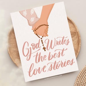 May include: A white rectangular print featuring an illustration of two hands holding a brown beaded rosary with a cross. The text "God Writes the best love stories" is written in a pink cursive script below the hands. The print has a textured finish.