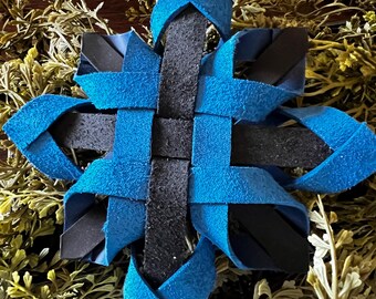 Puede incluir: Un adorno tejido en forma de estrella con un cuadrado negro central y extensiones azules en forma de pétalo. El adorno está hecho de material texturizado y descansa sobre un lecho de follaje verde y amarillo.