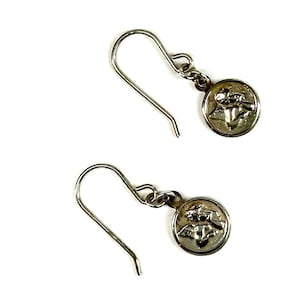 Puede incluir: Un par de pendientes colgantes plateados. Cada pendiente presenta un peque&ntilde;o dije redondo con un relieve detallado de un &aacute;ngel. Los pendientes tienen un dise&ntilde;o de gancho simple para orejas perforadas, sobre un fondo blanco.