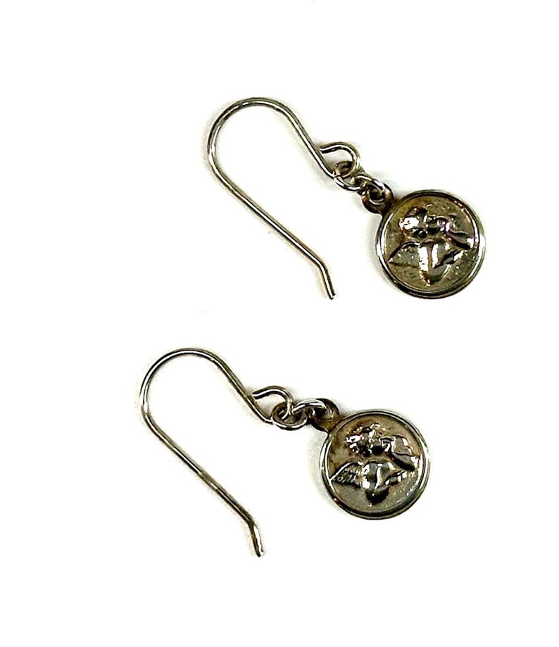 Puede incluir: Un par de pendientes colgantes plateados. Cada pendiente presenta un peque&ntilde;o dije redondo con un relieve detallado de un &aacute;ngel. Los pendientes tienen un dise&ntilde;o de gancho simple para orejas perforadas, sobre un fondo blanco.