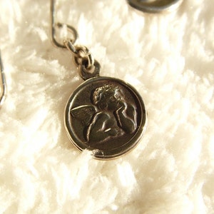 Puede incluir: Pendientes de &aacute;ngel plateados. Cada pendiente presenta un peque&ntilde;o colgante circular con un relieve detallado de un &aacute;ngel. Los pendientes tienen un dise&ntilde;o de gancho simple para orejas perforadas. El fondo es una superficie suave, blanca y texturizada.