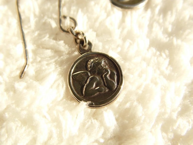 Puede incluir: Pendientes de &aacute;ngel plateados. Cada pendiente presenta un peque&ntilde;o colgante circular con un relieve detallado de un &aacute;ngel. Los pendientes tienen un dise&ntilde;o de gancho simple para orejas perforadas. El fondo es una superficie suave, blanca y texturizada.