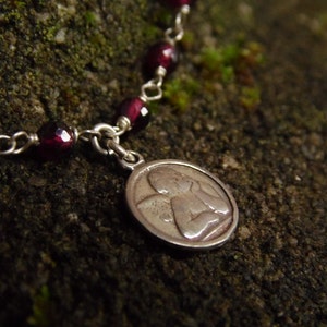 Puede incluir: Primer plano de una pulsera plateada con un colgante. La pulsera presenta cuentas facetadas de color rojo intenso y un peque&ntilde;o colgante ovalado con un dise&ntilde;o de &aacute;ngel grabado. Fondo texturizado verde oscuro.