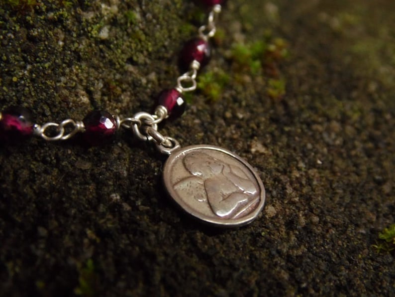 Puede incluir: Primer plano de una pulsera plateada con un colgante. La pulsera presenta cuentas facetadas de color rojo intenso y un peque&ntilde;o colgante ovalado con un dise&ntilde;o de &aacute;ngel grabado. Fondo texturizado verde oscuro.