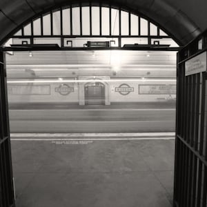 May include: A black and white photograph of a subway station entrance with a metal gate. The gate is open and the entrance is visible. The station is empty and the walls are tiled.