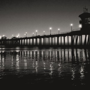 May include: A black and white photograph of a pier extending out into the ocean at night. The pier is lit by streetlights and the water is reflecting the lights.