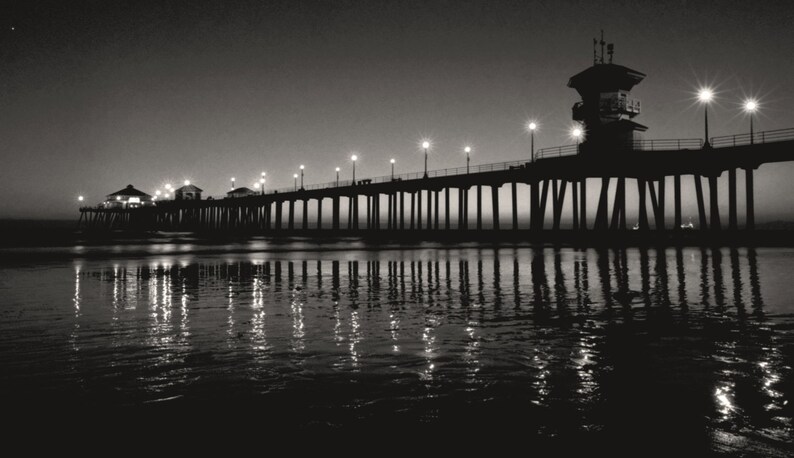 May include: A black and white photograph of a pier extending out into the ocean at night. The pier is lit by streetlights and the water is reflecting the lights.