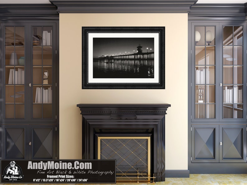 May include: Black and white photograph of a pier with lights at night. The pier extends into the ocean with a silhouette of the city in the background.