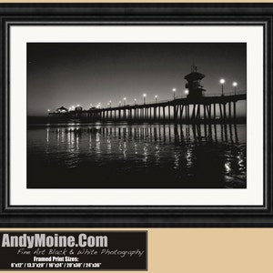 May include: A black and white photograph of a pier extending into the ocean at night. The pier is lit with streetlights and the water reflects the lights. The pier has a lifeguard tower at the end.