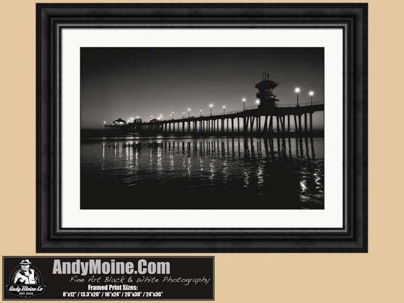 May include: A black and white photograph of a pier extending into the ocean at night. The pier is lit with streetlights and the water reflects the lights. The pier has a lifeguard tower at the end.