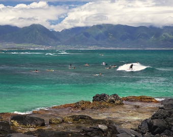 May include: A scenic view of a turquoise ocean with surfers riding waves. The ocean is surrounded by rocky shores and a mountain range in the distance.