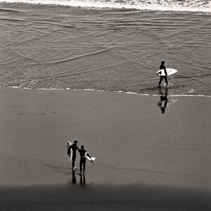 Op de afbeelding: Twee mensen in wetsuits lopen langs een zandstrand met surfplanken. Het strand is nat en de oceaan is op de achtergrond.