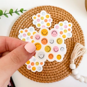 Puede incluir: Una mano sostiene una pegatina en forma de nube con caras sonrientes en naranja, rosa y amarillo. Pegatinas adicionales con dise&ntilde;os similares est&aacute;n sobre un posavasos tejido. El fondo es blanco.