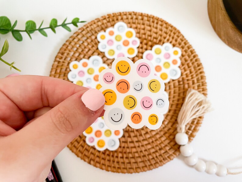 Puede incluir: Una mano sostiene una pegatina en forma de nube con caras sonrientes en naranja, rosa y amarillo. Pegatinas adicionales con dise&ntilde;os similares est&aacute;n sobre un posavasos tejido. El fondo es blanco.