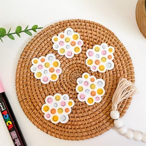 Puede incluir: Cinco pegatinas blancas con forma de nube, cada una cubierta con peque&ntilde;as caras sonrientes en amarillo, rosa, naranja y gris. Las pegatinas est&aacute;n dispuestas sobre un salvamanteles redondo y tejido. Tambi&eacute;n se ve un rotulador rosa y cuentas decorativas.