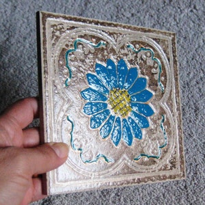May include: A square ceramic tile with a raised, textured surface. The tile features a blue and yellow flower design with intricate details.