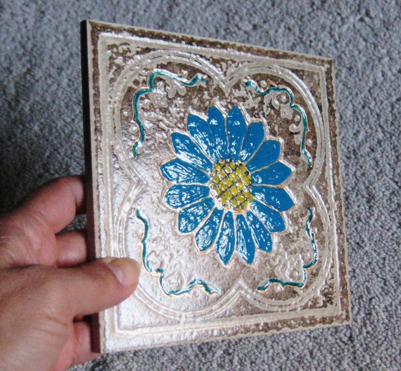 May include: A square ceramic tile with a raised, textured surface. The tile features a blue and yellow flower design with intricate details.