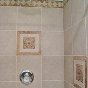 May include: A beige tiled shower cubicle with a chrome shower head and a chrome shower handle. The shower cubicle has decorative tile accents in the corners.