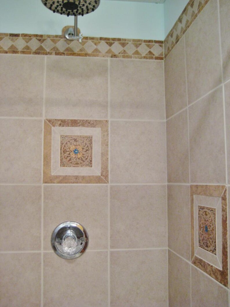 May include: A beige tiled shower cubicle with a chrome shower head and a chrome shower handle. The shower cubicle has decorative tile accents in the corners.