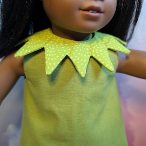 May include: A green doll dress with a distinctive collar. The dress is a solid green, and the collar is a lighter green with white polka dots. The dress is sleeveless and looks handmade.