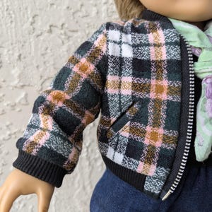 May include: A doll-sized bomber jacket with a plaid pattern in shades of green, pink, white, and brown. The jacket has a black ribbed collar, cuffs, and hem, and a full-length zipper. A small pocket is visible on the front.