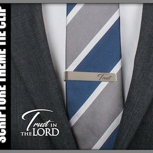 May include: A silver tie clip with the word "Trust" engraved on it. The tie clip is attached to a blue and gray striped tie.