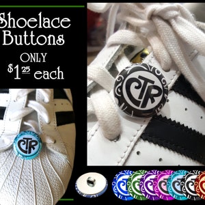 May include: Shoe lace buttons with a black and white design featuring the letters "CTR" in a circle. The buttons are shown on a white sneaker with black stripes. The buttons are available in a variety of colors, including green, purple, blue, and red.