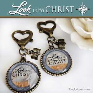 May include: Two gold heart-shaped keychains with a charm that says "Look unto Christ" in a blue and gold design. The charms are attached to a small book charm. The text on the charm reads "Doubt not, fear not".