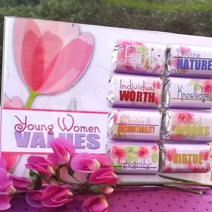 Peut inclure: Une boîte en acrylique transparente avec un fond violet et un motif de tulipe rose en aquarelle. La boîte contient huit petites barres de chocolat enveloppées dans du papier d'aluminium avec des motifs floraux et du texte qui dit "Foi", "Nature Divine", "Valeur Individuelle", "Connaissance", "Choix et Responsabilité", "Bonnes Œuvres", "Intégrité" et "Vertu". Le texte "Valeurs des Jeunes Femmes" est imprimé sur la boîte.