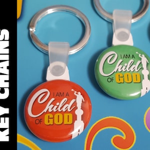 May include: Two keychains with a silver key ring and a round button. The buttons are green and orange and have the text "I am a Child of God" in white and yellow lettering. The buttons have a silhouette of a person holding a trumpet.