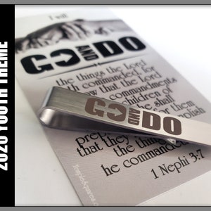 May include: A silver metal tie clip with the words "GO and DO" engraved in brown. The tie clip is on top of a card with the text "GO and DO the things the Lord hath commanded, for know that the Lord commanded his children to prepare that they might do the things which he commanded." 1 Nephi 3:7.