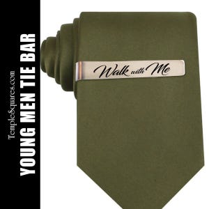 May include: An olive green tie with a silver tie bar. The tie bar is engraved with the words "Walk with Me" in a cursive font. The text "YOUNG MEN TIE BAR" is printed vertically on the left side of the image.