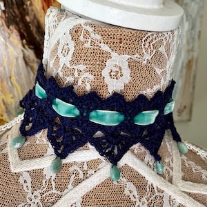 May include: A navy blue crochet choker necklace with a scalloped edge. The necklace features teal velvet ribbon accents and dangling teardrop-shaped beads. The choker is displayed on a mannequin.