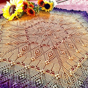 May include: A handmade, circular crochet doily in shades of tan, transitioning to purple at the edges. The intricate design features diamond and floral patterns. Yellow sunflowers and orange accent flowers are arranged on the top.