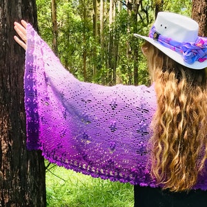 Pode incluir: Um xale de crochê roxo e lavanda com um delicado padrão de renda. O xale está drapeado sobre o braço de uma pessoa e é mantido no lugar por sua mão. O xale está em um ambiente natural com árvores ao fundo.
