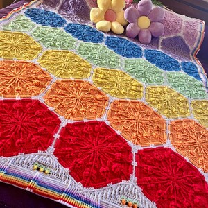 May include: A colourful, handmade crochet blanket with a hexagonal pattern. The blanket displays a vibrant spectrum of colours, including red, orange, yellow, green, blue, and purple. Two flower-shaped cushions rest on top of the blanket.