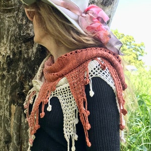 May include: A woman wearing a white hat with a pink and orange scarf, a black sweater, and a crocheted shawl with brown and cream accents.