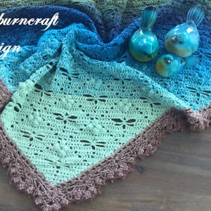 May include: A crocheted shawl in shades of blue, green, and brown with a delicate lace border. The shawl is folded and draped over a wooden surface with decorative glass birds.