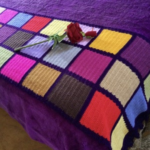 May include: A colorful crocheted throw blanket with a square pattern in shades of pink, purple, yellow, blue, brown, and red. The blanket is on a purple bedspread and has a single rose on top.