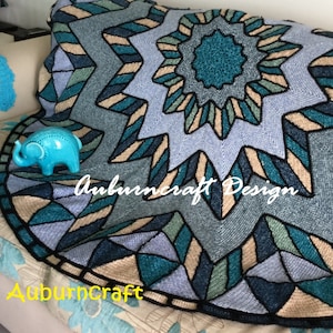 May include: A crocheted blanket with a large, geometric star design in shades of blue, teal, and beige. The blanket is made with a black border and has the text "Auburncraft Design" in the center.