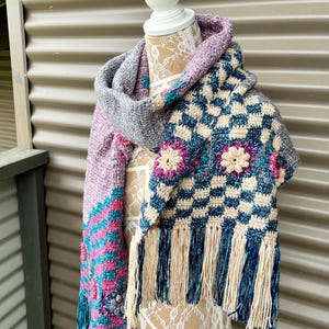 Puede incluir: Una bufanda de ganchillo con un patrón de cuadros y detalles florales. La bufanda está hecha con una variedad de colores, incluyendo rosa, azul y gris. Tiene flecos en los extremos.