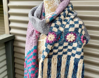 May include: A crocheted scarf with a checkered pattern and floral accents. The scarf is made with a variety of colors, including pink, blue, and gray. It has fringe on the ends.