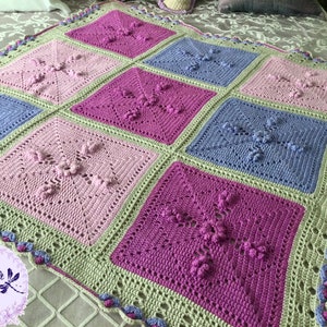 May include: A crocheted baby blanket with a square pattern in shades of pink, purple, green, and blue. The blanket has a scalloped border.