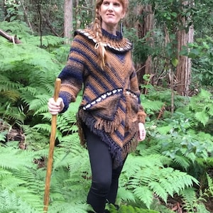 May include: A brown and blue striped crocheted poncho with fringe. The poncho has a high neck and a white crocheted trim. The model is wearing black leggings and is standing in a forest.