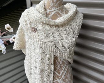 May include: A white crocheted shawl with a textured pattern and a small decorative pin. The shawl is draped over a white mannequin.