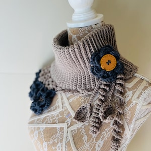 May include: A hand-crocheted cowl in taupe and blue, decorated with a blue flower and a mustard button. The cowl has a ribbed texture and decorative crochet details. A stylish accessory to add a touch of handmade charm to any outfit.