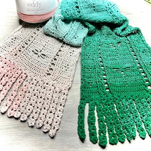 Peut inclure: Deux écharpes crochetées à la main dans les tons de vert, bleu et beige avec des franges. L'une est un dégradé de vert, l'autre de beige à rose. Une pelote de laine rose est dans le coin supérieur gauche.