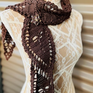 May include: A dark brown crocheted scarf featuring an openwork pattern and a decorative flower-shaped brooch with light-colored petals attached to the fabric. The scarf is draped over a mannequin torso covered in white lace fabric.